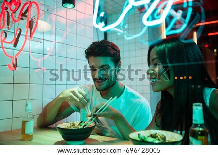 Picture of young multiethnic smiling loving couple sitting in cafe looking aside eating.
