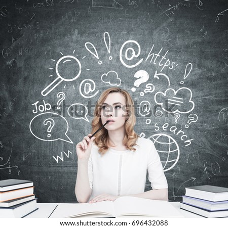 Beautiful young woman is sitting at a table with piles of books on it. The one in front of her is open. She is thinking and biting a pencil. Blackboard with an internet search sketch