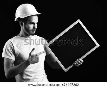 handsome bearded builder or foreman in white shirt and helmet on serious face holding board on black studio background, copy space