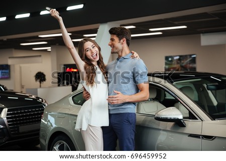 Picture of young loving couple standing near car in car dealership. Looking camera showing keys make winner gesture.