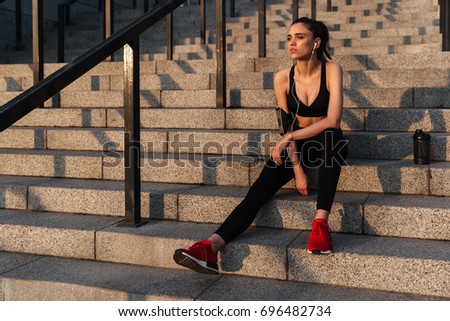 Picture of serious young sports lady sitting with earphones outdoors and looking aside.