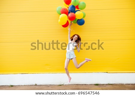 Picture of young smiling redhead woman jumping over yellow wall. Have fun with balloons.