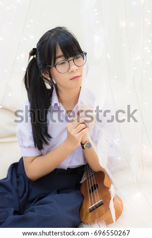 Cute Asian Thai high school girl in uniform and glasses sitting and holding a soft cloth curtain decorate with feather and light with guitar by her side in schoolgirl fashion music recreation concept
