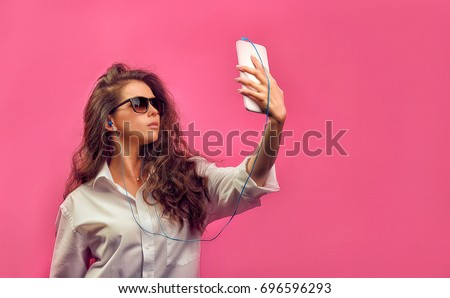 Beautiful caucasian woman in a white shirt  in glasses with headphones, holding a white tablet in hands and makes selfie photo on a pink bright  background. Studio photo with a stylish model.