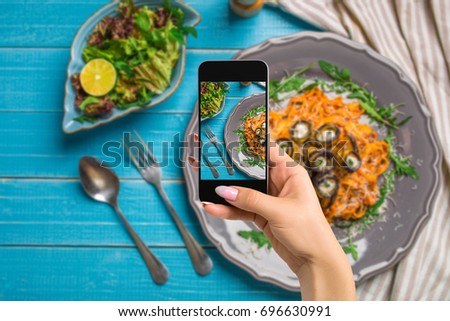 Photographing food concept - woman takes picture of pasta with eggplants, tomato, cheese, arugula and salad