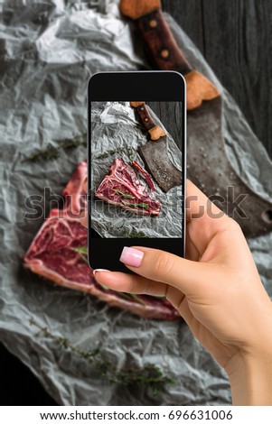Photographing food concept - woman takes picture of raw dry aged t-bone steaks for grill with fresh herbs and cleaver