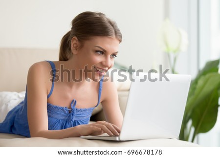 Happy smiling young woman using laptop while resting on sofa at home. Beautiful lady relaxing and having fun with electronic device, playing computer games, chatting with friends online, enjoying app