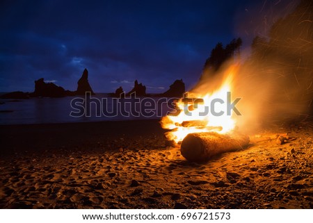 Stack of wood set on fire on a beautiful sandy shore with a surreal rocky formation in the background. Picture taken in Shi Shi Beach, Neah Bay, Washington Coast, USA, at night after a cloudy sunset.
