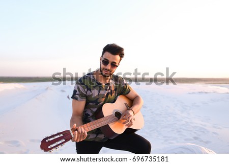 Handsome Arabian guy plays tune on guitar, drives strings and sings song, sitting on hill in middle of wide sandy desert on warm summer evening at sunset. Swarthy man with dark hair and short haircut