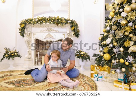 Cute little girl child holding gadget and plays game running his finger along touchscreen via his father, handsome man who told girl how to use tablet and sitting on background of fireplace with