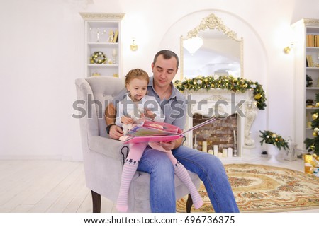 Cute blond baby with his father reading pink baby big book. man tickles and kisses his daughter, laughing girl sitting on lap of pope in gray big chair on background of fireplace with candles