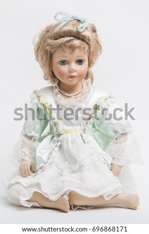 Portrait of a sitting ceramic porcelain handmade vintage doll with long blond hair and elegant hairstyle in an old linen dress with floral decorative embroidery on white background.