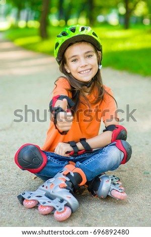 Happy girl in a protective helmet and protective pads for roller skating sitting on the road and showing thumbs up