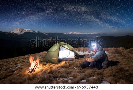 Male tourist have a rest in his camp at night. Man with lighting headlamp sitting near campfire and tent, looking to the camera under beautiful sky full of stars and milky way Royalty-Free Stock Photo #696957448