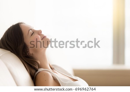 Relaxed young woman enjoying rest on comfortable sofa, calm attractive girl relaxing on couch, breathing fresh air with eyes closed, meditating at home, peace of mind, headshot portrait, copy space Royalty-Free Stock Photo #696962764