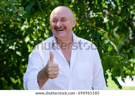 Successful excited elderly man in white shirt showing ok sign while standing in a garden background. 