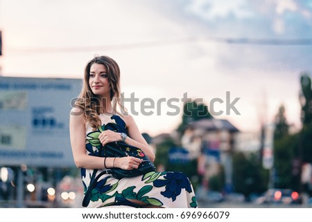 Fashionable girl sitting on handrail. Keeping a designed backpack on the handrail. Looking for old textbooks inside backpack. Big buildings in the background. Blonde hair putting forward.