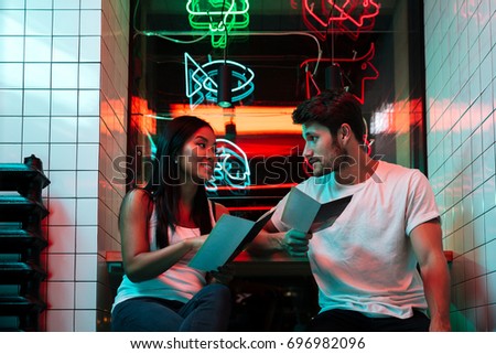 Picture of young multiethnic happy loving couple sitting in cafe looking aside holding restaurant menu.