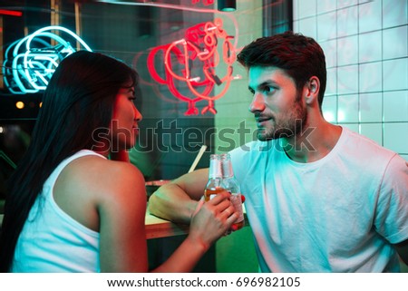 Picture of young multiethnic cheerful loving couple sitting in cafe looking aside drinking beer.