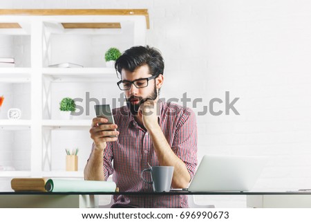 Portrait of attractive young businessman using cellular phone while sitting at office desk with laptop and other items. Leisure, break, communication and technology concept