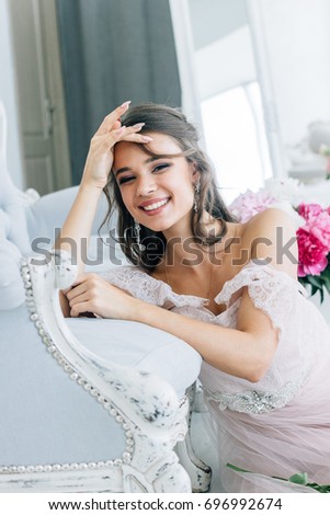 Beautiful bride in a white dress is sitting near the couch and smiling. In the background is a beautiful bouquet