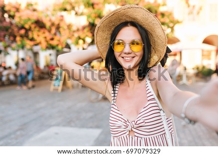 Cheerful girl holding hat while making selfie and laughing waiting friends on main city square. Close-up portrait of smiling young lady in glasses with black straight hair taking picture of herself.