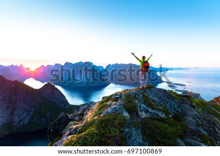 Happy woman hiker enjoying scenic view of midnight sun at the top of Reinebringen hike above Reine village in the Lofoten archipelago during arctic summer, Norway Royalty-Free Stock Photo #697100869