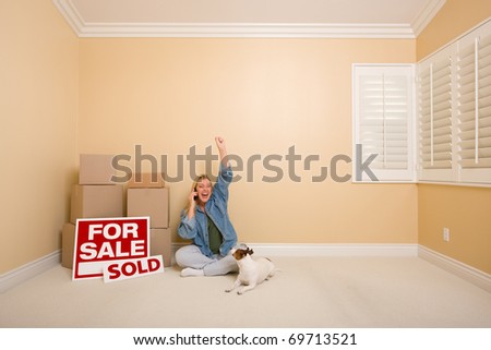 Pretty Woman Sitting on Floor Using Phone Celebrating Next to Moving Boxes, Sold Real Estate Signs and Dog in Empty Room.