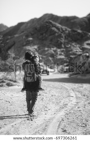 Father carrying his tired after trip son in his arms. Boy holding his favorite teddy bear toy in his hands. Black and white road & mountains landscape. Outdoor quality time. Parenting 