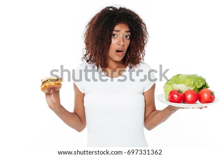 Picture of confused young african woman standing isolated over white background. Choosing between burger and vegetables.