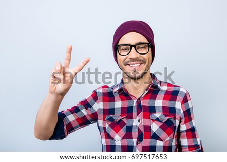 Peace! Smiling young nerdy stylish student hipster is gesturing vsign on pure background in black trendy glasses and hat, casual bright checkered shirt