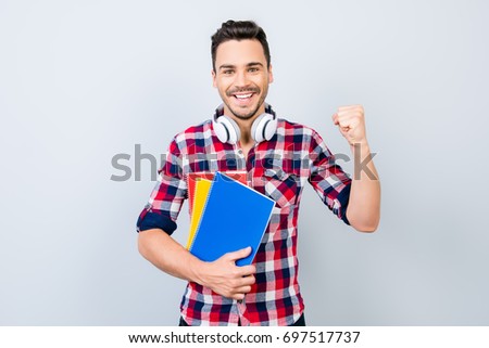 Cheerful young brunet nerdy student with stubble is standing with colorful books on pure background in casual checkered outfit, whitemodern big headphones. He is gesturing victory, he passed exams