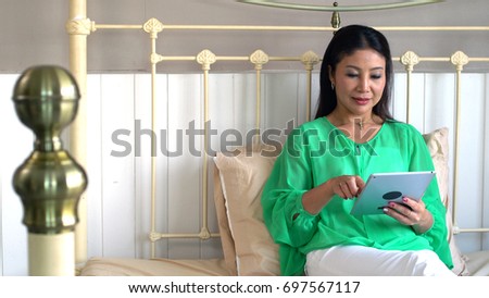 The Asian beautiful mother use tablet on the bed and lovely teenage daughter bring hot black coffee to her mom, they have activities together joyful in bedroom, teenage family relation concept.
