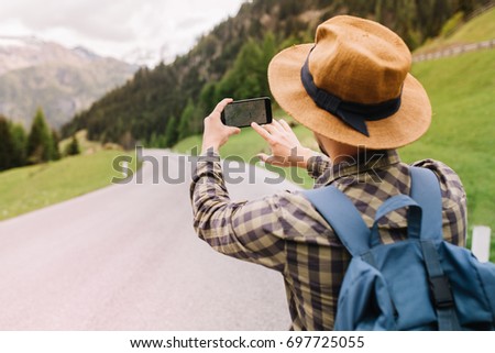 Male traveler in brown hat searching right road by using online map standing on beautiful mountain background. Outdoor portrait of walking young man with backpack having fun in weekend.