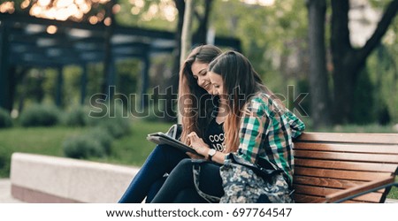 MODERN GIRLS SITTING ON A STYLISH WOODEN BENCH. BACK TO SCHOOL LIFE WITH FRIEND. FRIENDSHIP GOALS. SHOWING OLD PHOTOS OF SCHOOL LIFE ON tablet AND SMILING AT IT. SCHOOL BACKPACK ON THE BENCH.