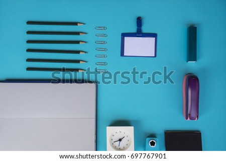 Flat lay work desk accessories, computer laptop or notebook, calculator,card holder, paper puncher, clock,  pencil, and paper clip on solid blue background.