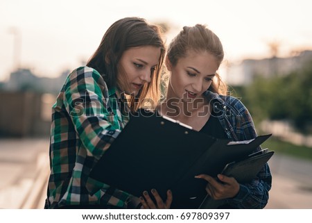 Girl showing her folder her friend and both bursting out a laugh. Going through some funny mistakes Girls looking pretty cute on laughing. Beautiful city center on background. Students back to school