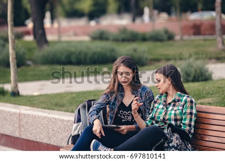 Modern girls sitting on a wooden bench. Talking seriously about their school supplies. Worried about the school supplies list. Looking at her sneakers. School backpacks are on bench. Back to school
