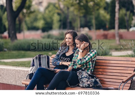 Modern girls sitting on a wooden bench. Talking seriously about their school supplies. Worried about the school supplies list. Looking at her sneakers. School backpacks are on bench. Back to school