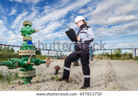 Woman engineer in the oilfield talking on the radio wearing white helmet and work clothes. Industrial site background. Oil and gas concept.