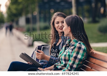 Teen girls wandering around the park. Taking rest on a wooden bench. Talking about their school supplies list. Anxious to hear what she is telling. Pleasant atmosphere and messy hair. Back to school
