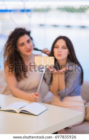 Two young beautiful women sit outdoor and do selfie-photo on a phone, one of them smiles, another gives air kiss