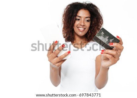 Picture of cheerful young african lady standing isolated over white background. Looking aside using mobile phone holding credit card.