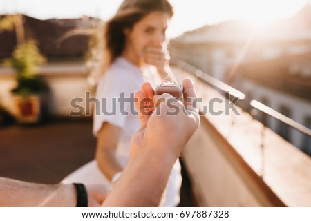 Man holding wedding ring in front of astonished happy girl covering mouth with hand. Romantic photo of charming woman standing on roof early in evening on date with boyfriend in anniversary. Royalty-Free Stock Photo #697887328
