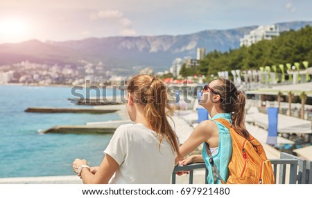 Two young women, laughing girl with small orange backpack and her girlfriend stands on the pier on background of sea