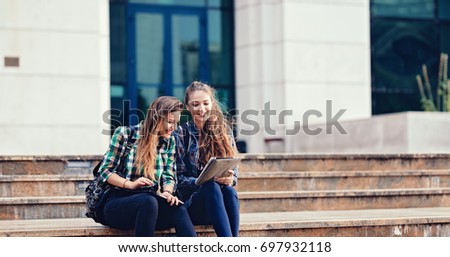 Stylish girls standing in front of school in pretty outfits and make up Showing something interesting in the tablet to her friend the pictures of her vacation tour and grinning Students back to school