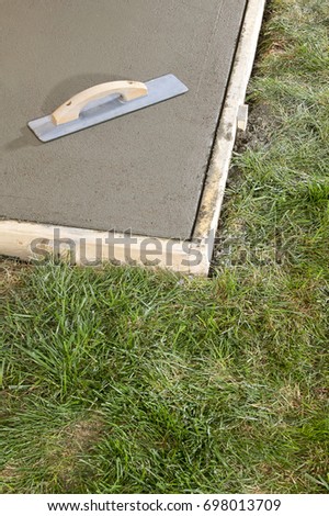 Angled view of a trowel tool sitting on top of wet liquid cement in a wooden frame surrounded by grass