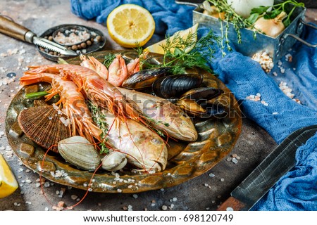 Fresh sea fish, prawns, clams on plate at stone table with knives, lemon