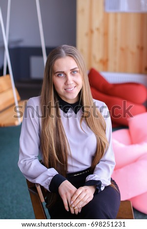 Portrait of cute long-haired successful secretary girl who poses on camera after end of day and smiles in modern office in afternoon. Woman with fair-haired hair dressed in light blouse with black