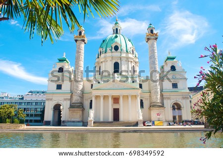 Facade of Karlskirche (St. Charles's Church) against blue sky, white clouds and a pond with water on a hot summer day. Green tree branch with pink flowers on the front. Vienna, Austria
